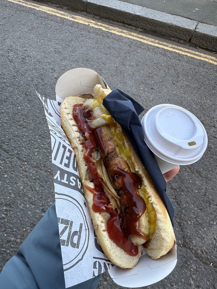 A hot dog with ketchup and mustard, held beside a takeaway coffee